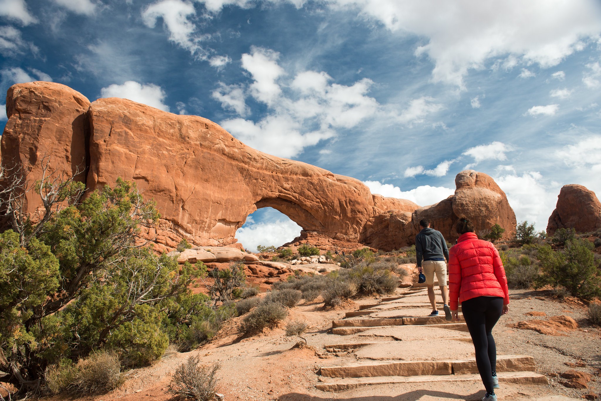 Two people, one in a red coat and black pants and one in a navy shirt and tan pants, walk up a gravel trail surrounded by green trees towards a large red natural stone opening with blue sky and white clouds behind.