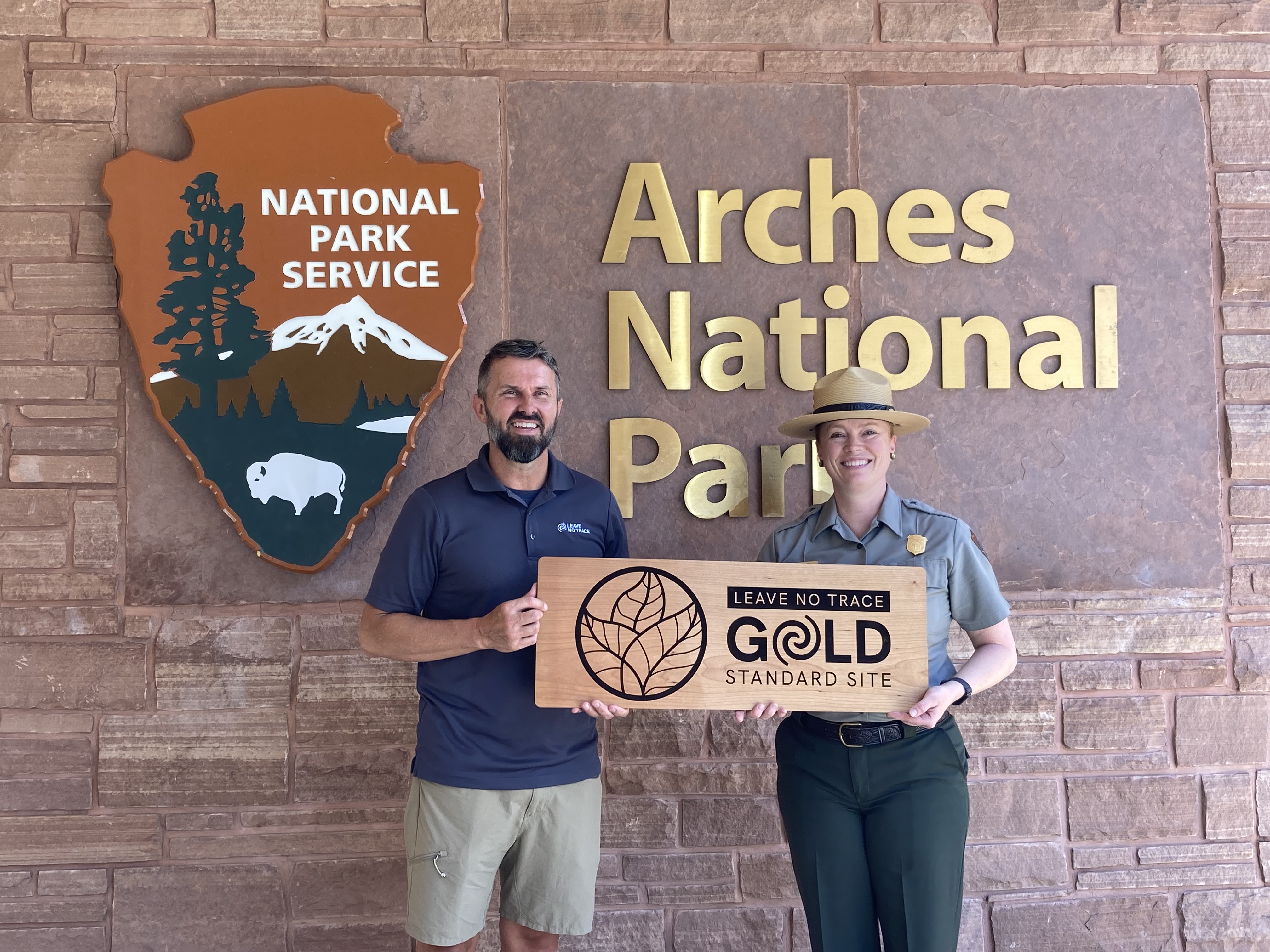 two people hold engraved wooden plaque in front of wall with NPS arrowhead and gold text reading "Arches National Park" two people hold engraved wooden plaque in front of wall with NPS arrowhead and gold text reading "Arches National Park"