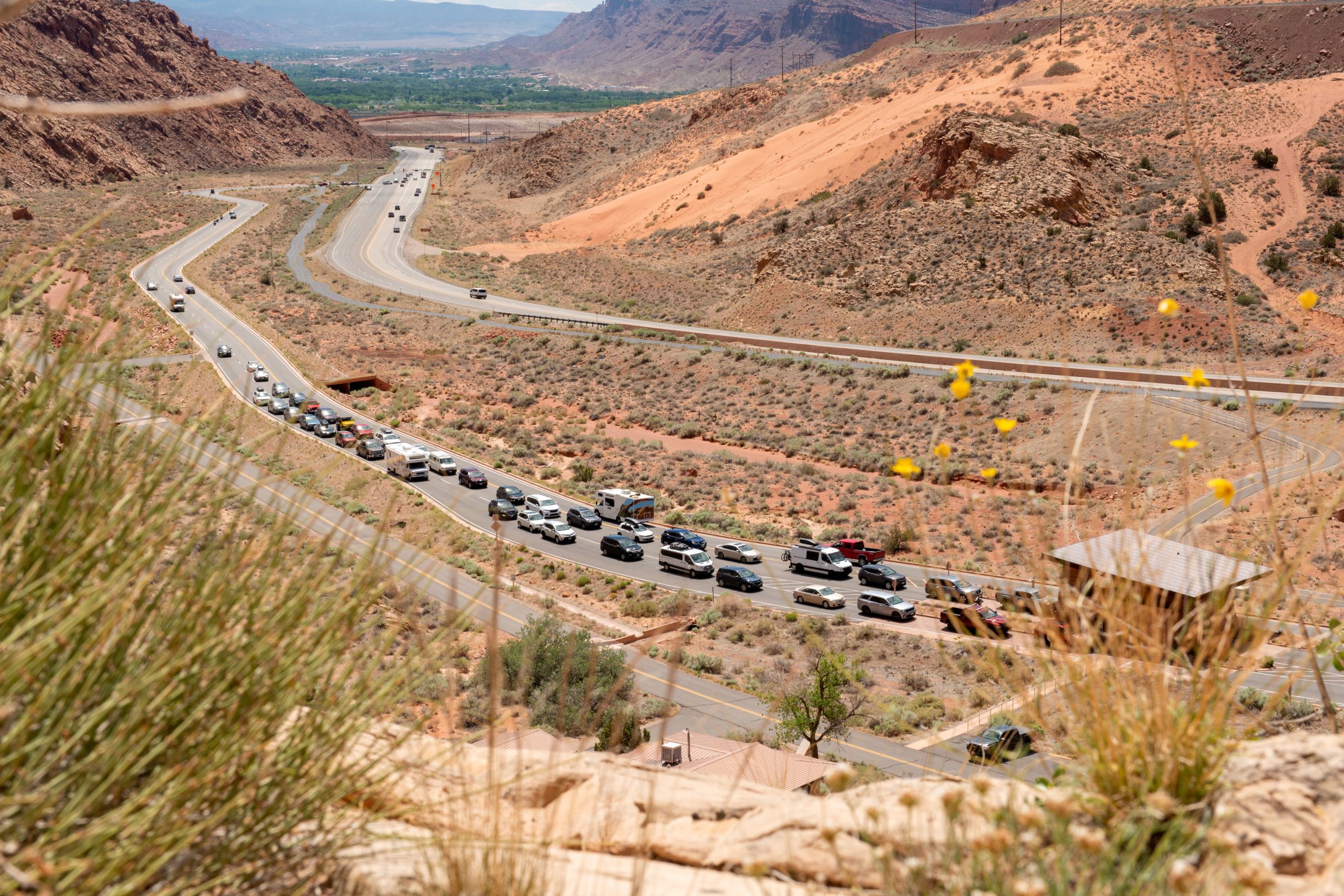 A long line of vehicles of various colors and sizes approach the Arches entrance station. Yellow flowers and tan cliffs rest in the foreground. A long line of vehicles of various colors and sizes approach the Arches entrance station. Yellow flowers and orange cliffs rest in the foreground.