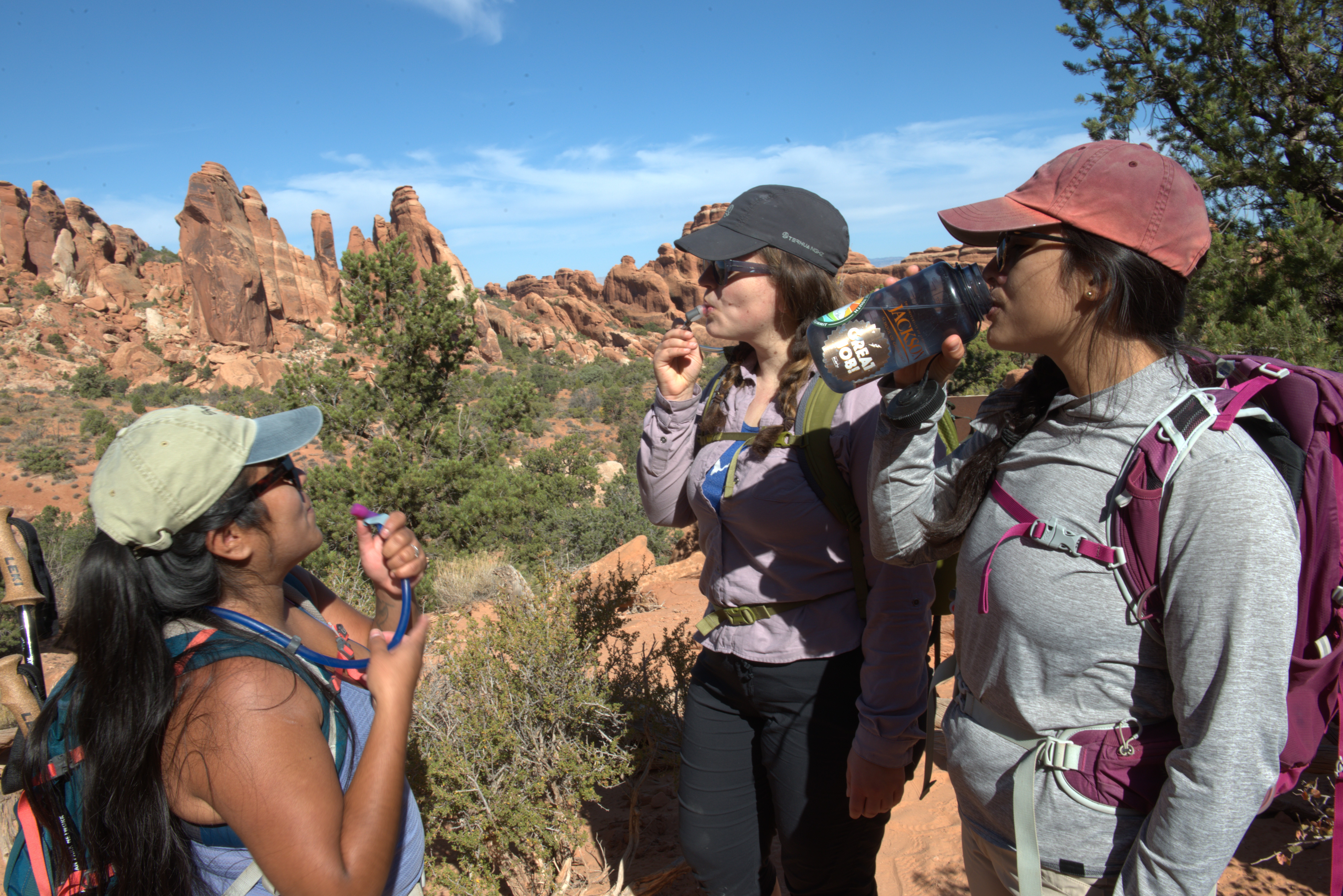 Three people wearing hiking clothing and backpacks drink water along a trail. Desert landscape with sandstone spires are behind them.