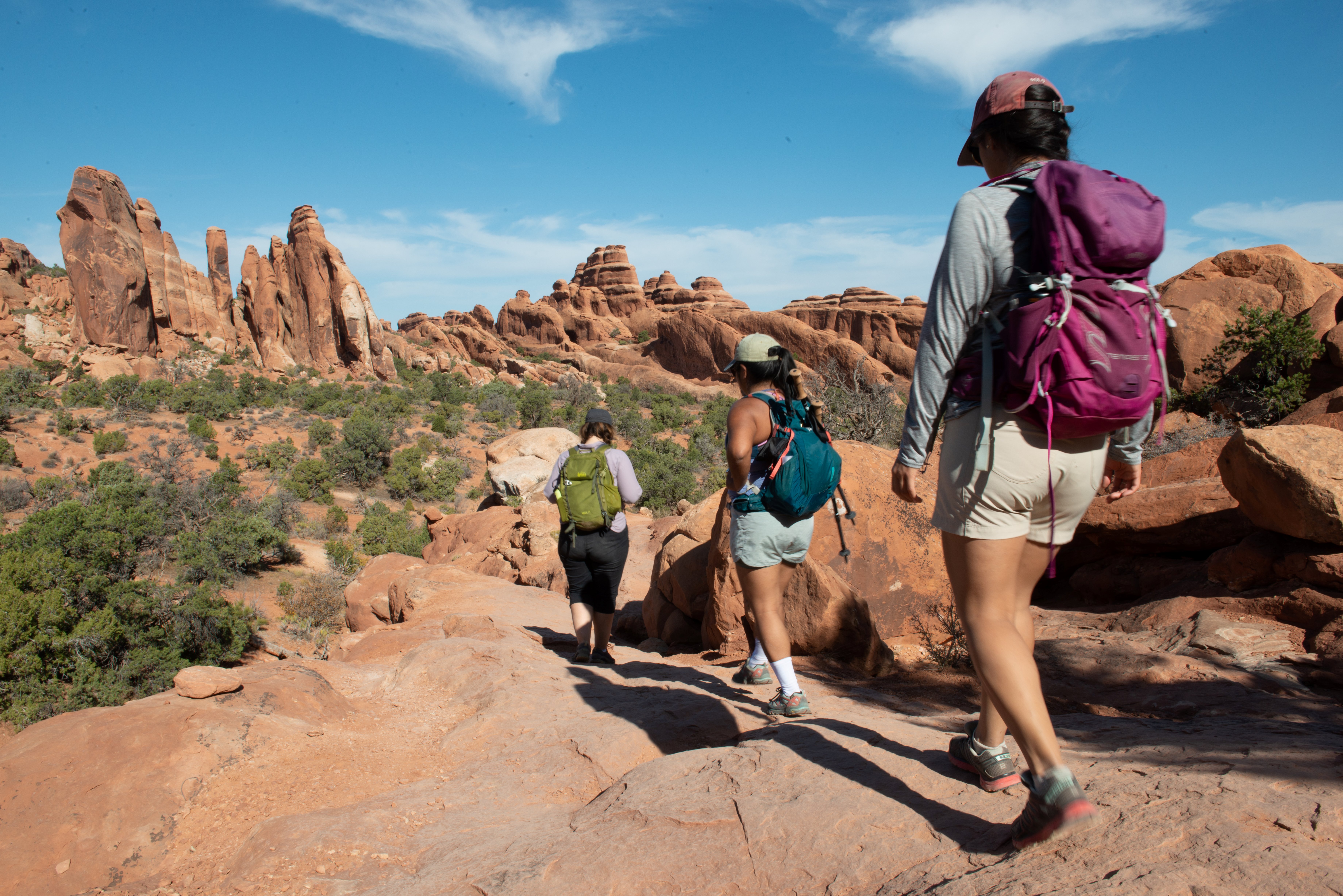 three hikers on rocky trail, desert shrubs and tall sandsone fins in background three hikers on rocky trail, desert shrubs and tall sandsone fins in background