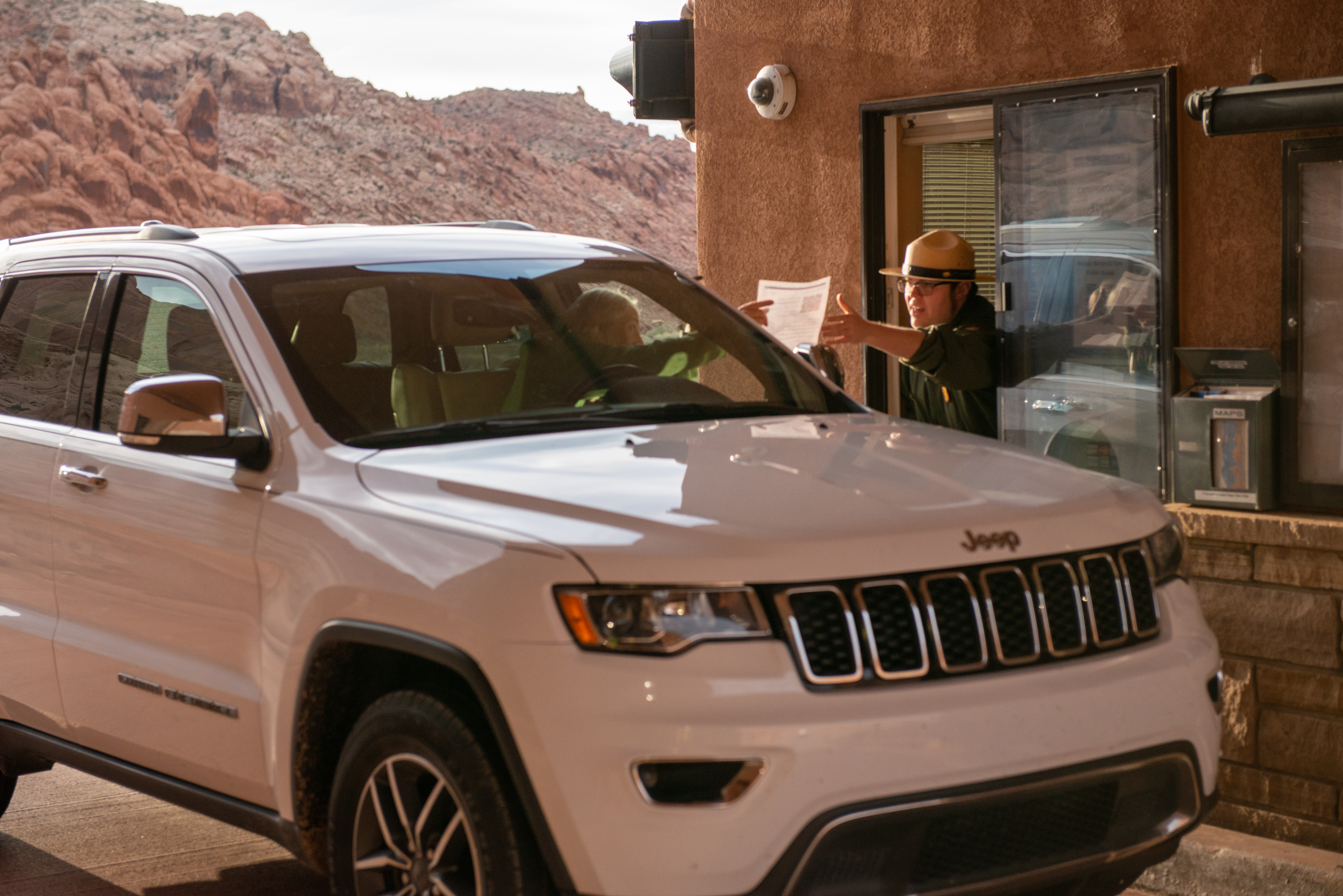 person driving white jeep hands paper to park ranger leaning out of booth window