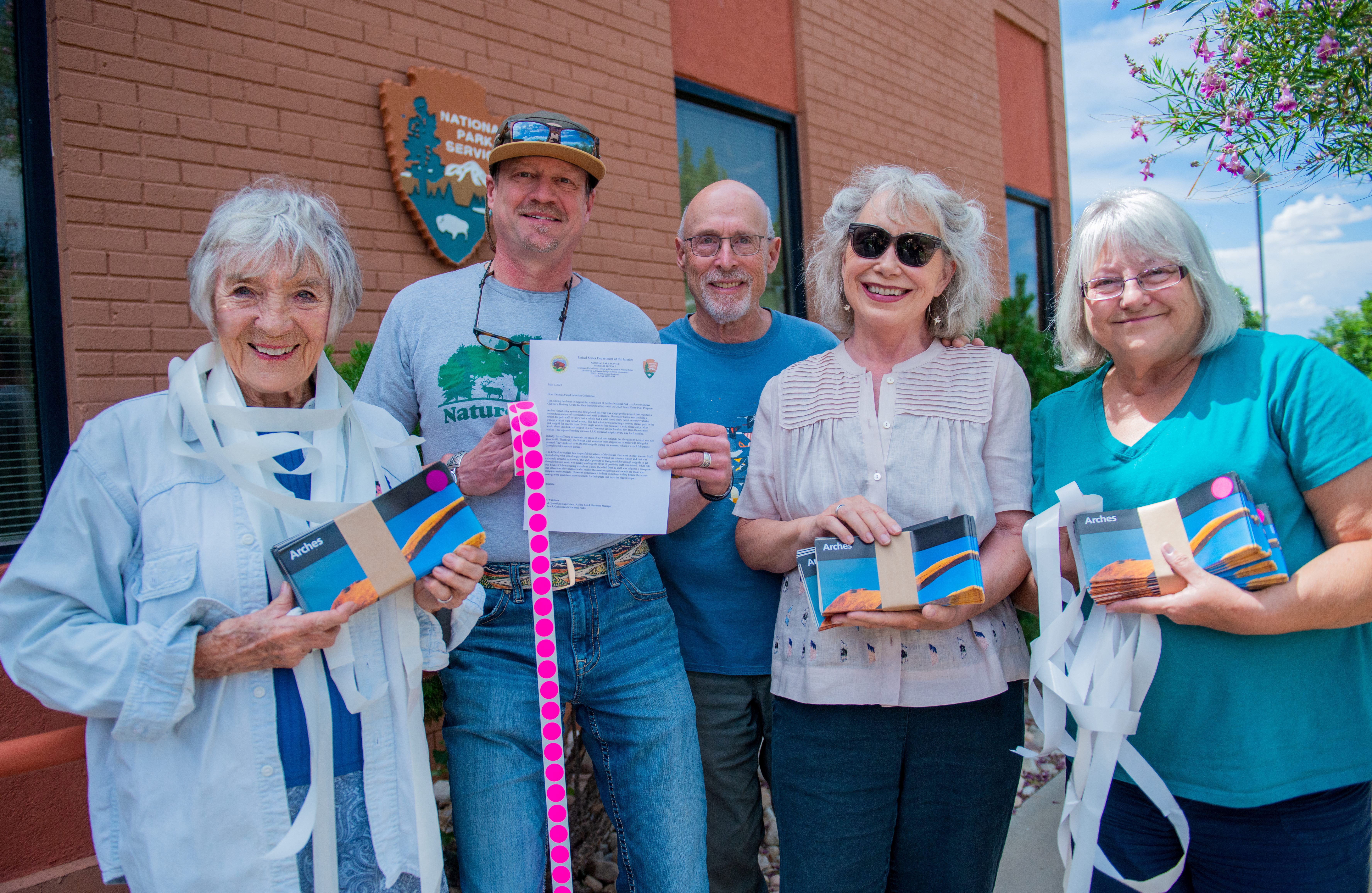 Five standing seniors hold packs of Arches maps and a roll of pink circle stickers. Five seniors stand outside a building, holding Arches maps and rolls of pink stickers