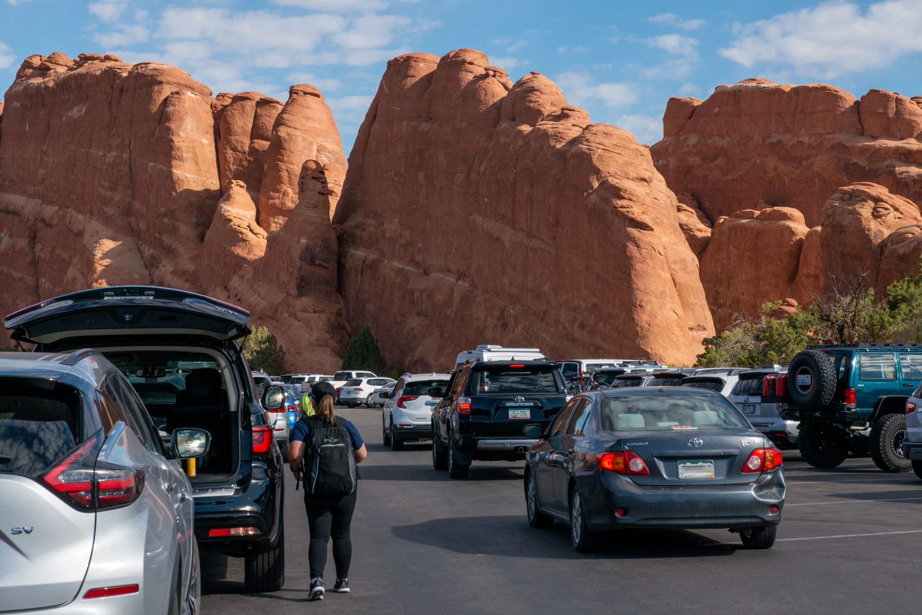 A woman walks along the edge of the Devils Garden Parking lot next to many different vehicles and large, orange, sandstone fins.