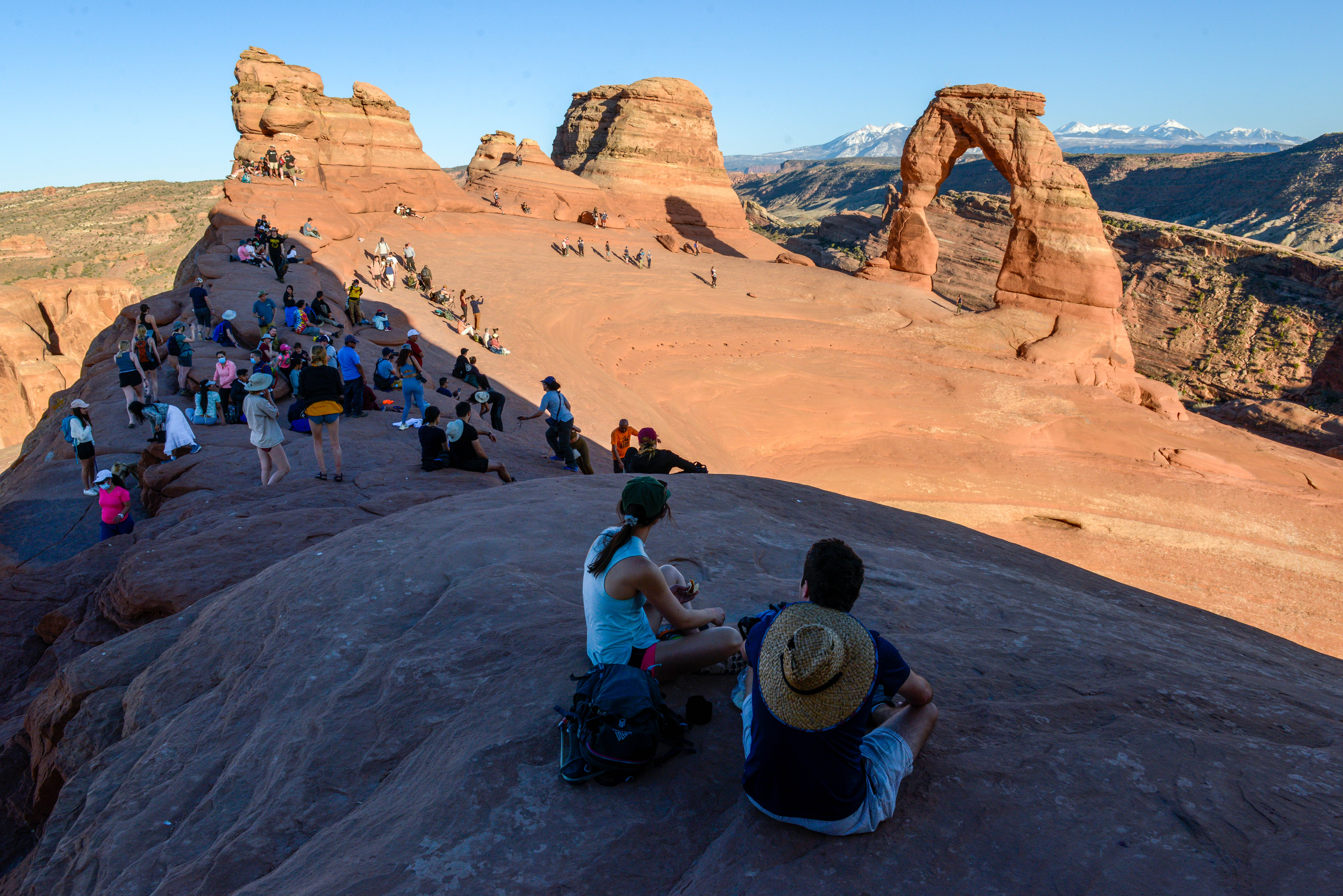 Several dozen visitors sit or stand in front of Delicate Arch, an orange sandstone arch perched on the precipice of a cliff framing mountains in the distance.