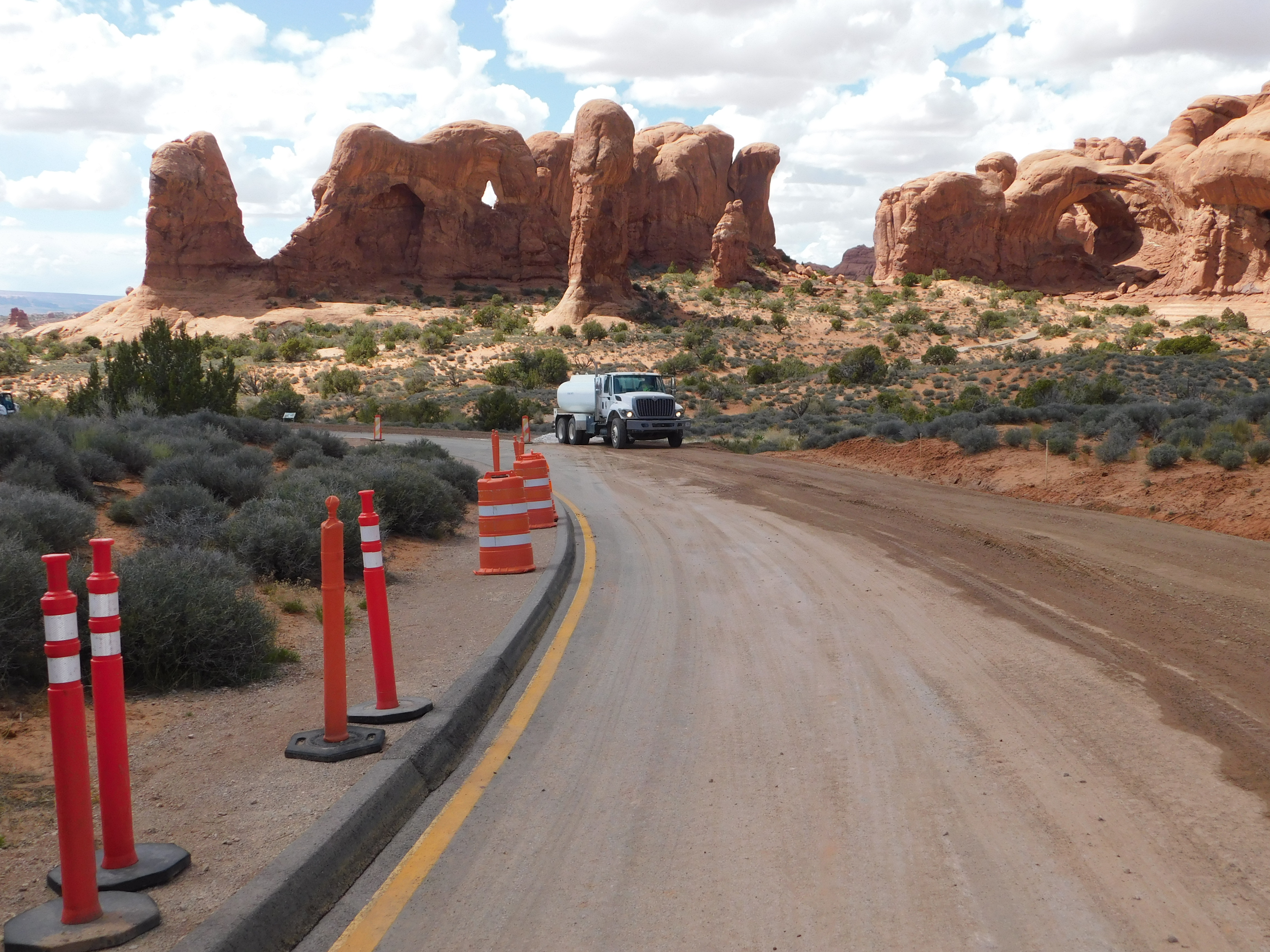 Orange cones and a large work truck sit along a curved road with two large sandstone arches in the background.