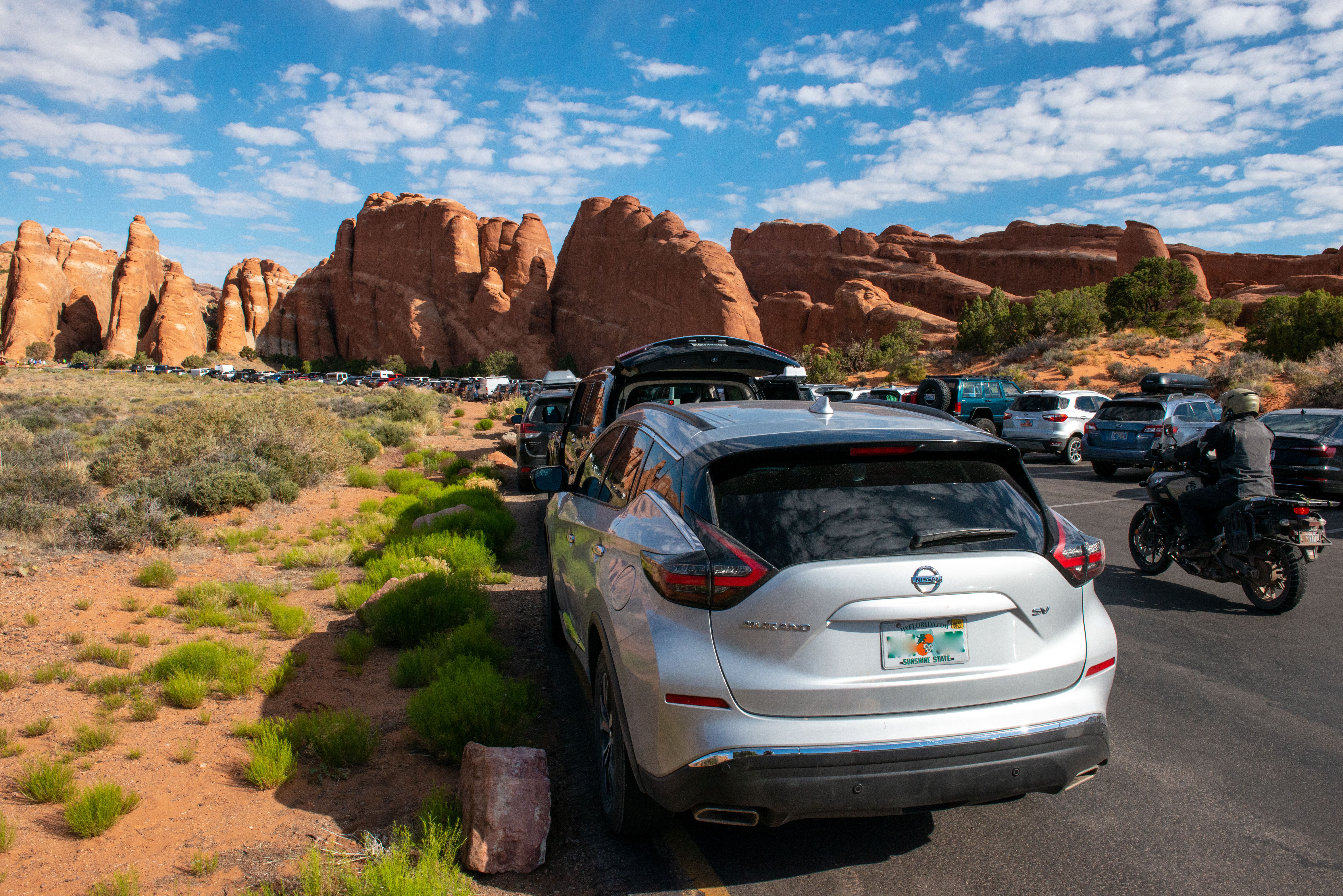 Cars line the parking lot of Devils Garden on a sunny day. A motorcycle passes by. In the background, orange sandstone spires and formations are visible.