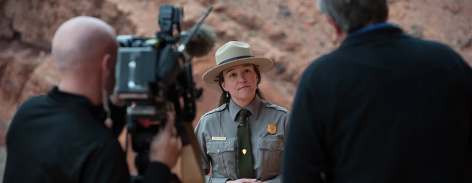NPS/Neal Herbert A female park ranger stands in front of a video camera surrounded by steep red rock walls.