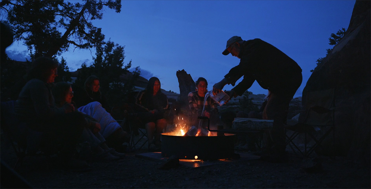 People gather around a campfire at dusk while a person dumps water on it to drown the flames