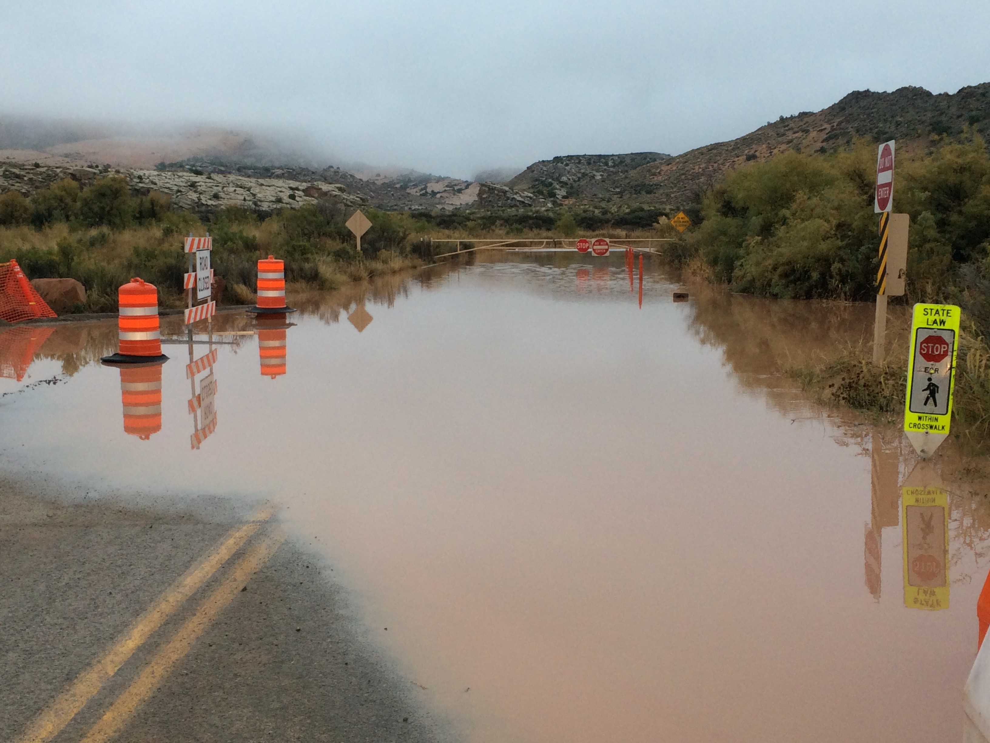brown water covers a paved roadway for 50 feet in front of a barricade, low shrubs on roadsides brown water covers a paved roadway for 50 feet in front of a barricade, low shrubs on roadsides