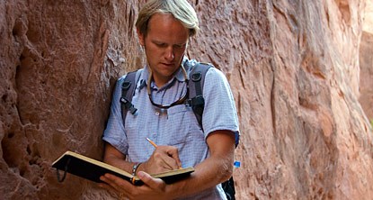 A man leaning against a rock wall writing in a notebook.