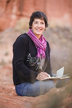 A woman sits in red rock landscape holding a pen to an open notebook.