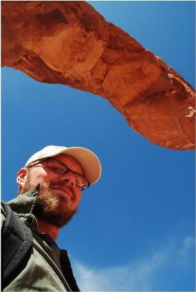 Man in a ballcap with eyeglasses on, standing under an arch, looking down at the camera.