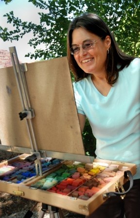 A woman in nature standing behind an easel with pastels arranged in front of her.