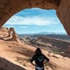 A person stands beneath an arch looking out at a vast landscape of sandstone rock features and mountains in the distance.