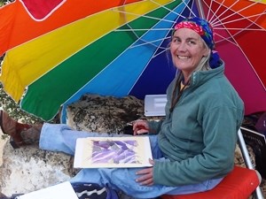A woman seated beneath rainbow umbrella with a painting in her lap.