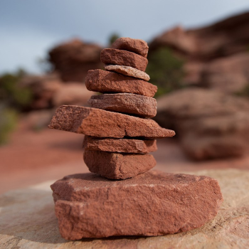 A stack of tan rocks against a background of red rocks, green plants, and blue sky.