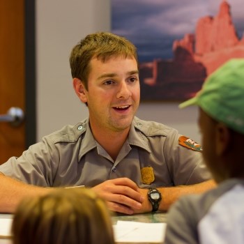 A male ranger in uniform behind a desk talking to children.