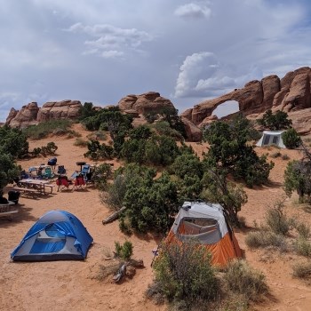 A blue tent, orange tent, and white tent sit in sand between green trees at a campsite with a fire rings, picnic tables, and camp chairs. In the background is an orange colored natural stone arch, with blue sky and white clouds behind.