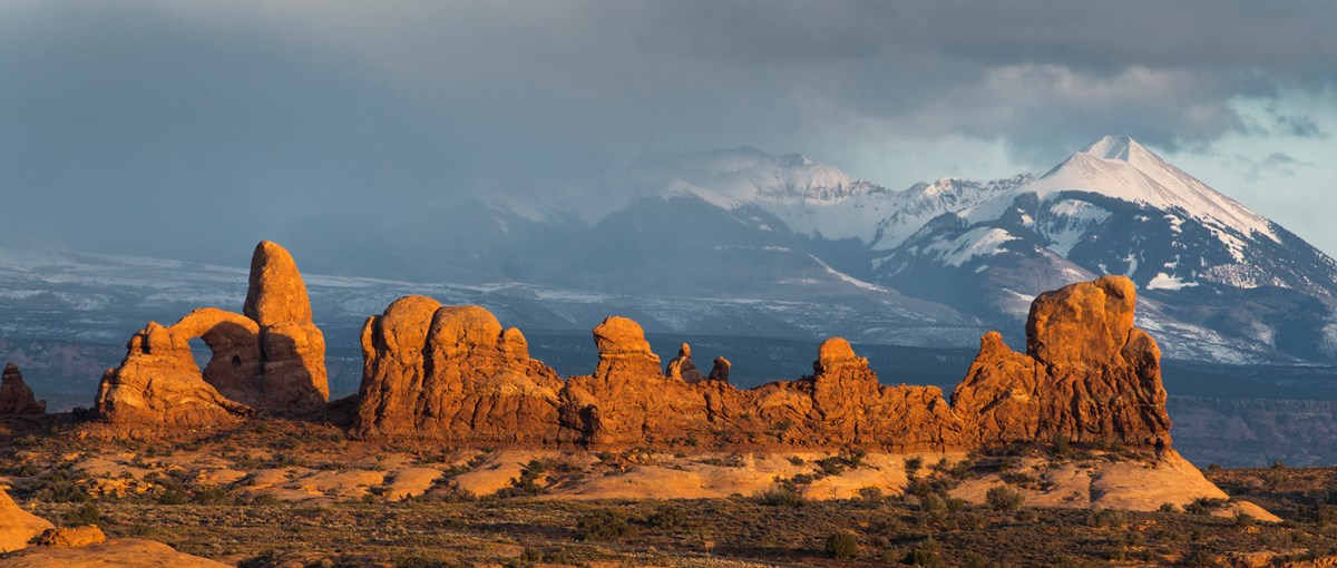 Towering peaks of red rock with an arch, being illuminated by a low sun. The scene is backdropped by cloud covered snowy capped mountains,