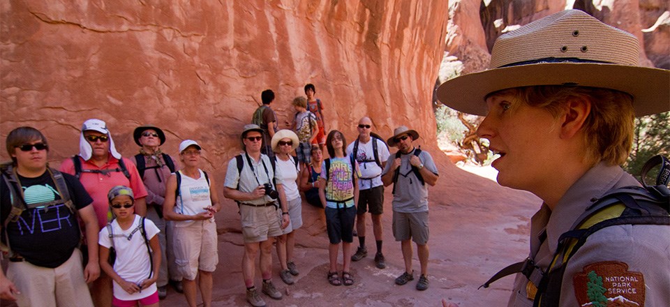 A ranger is talking with a group of people on a guided hike. They are standing in the shade of a large red rock wall.