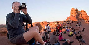 A man has a camera raised to his eye to take a photograph of an out of frame view. Behind him is an expanse of bare red rock is covered by people. The low light of the sunset is causing the rocks to be illuminated in a warm glow.