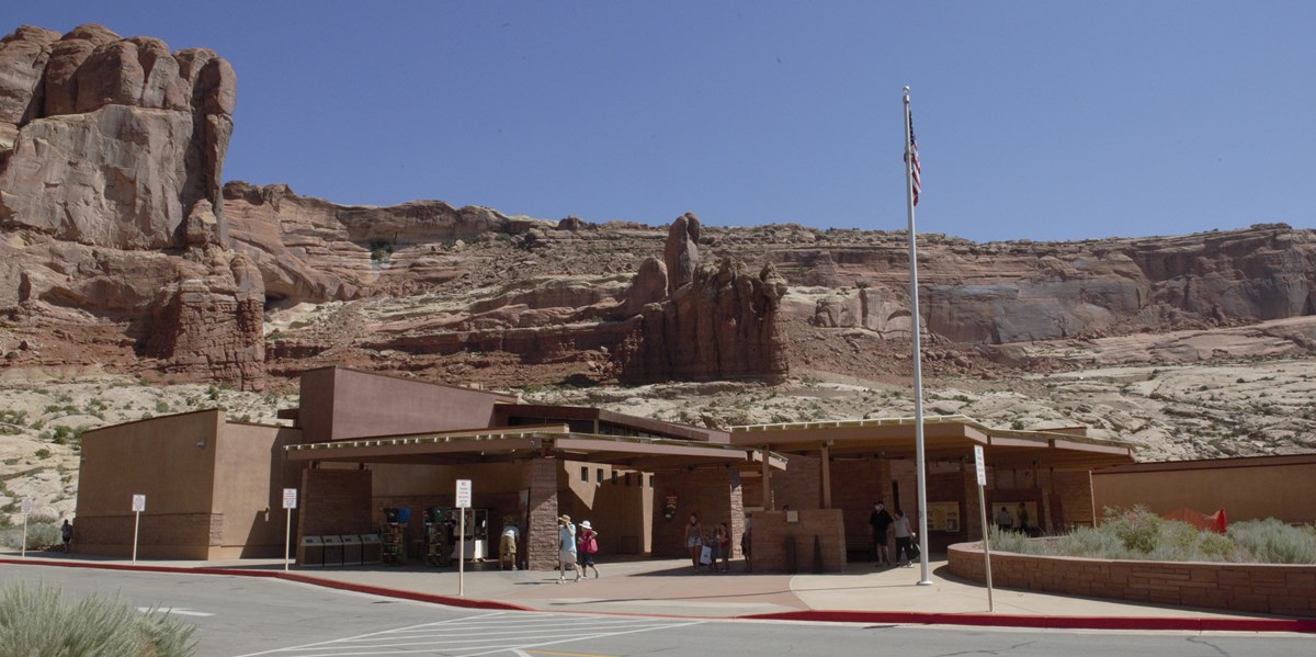 A large single story sand colored building with people walking around outside. The American flag is hung on a flagpole. Behind the building are massive red rock cliffs and a bright blue sky.