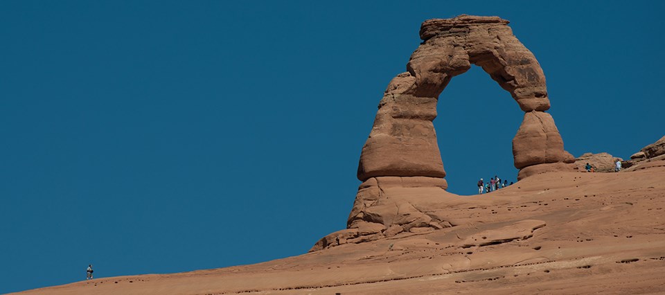 A large sloping red rock face with a unique rock formation jutting out of it. It is an arch shaped like an upside U. The blue sky can be seen through the wide opening in the rock. The people standing below it look very small.