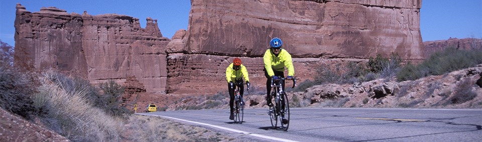 two cyclists in bright yellow jackets ride on a paved road two cyclists in bright yellow jackets ride on a paved road