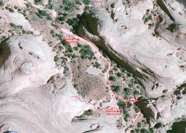 Aerial view of a narrow canyon with a red line drawn to show route. There is bare sand colored rock on either side with a dry sandy wash surrounded by green vegetation.