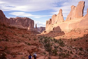 Two hikers walk down a trail through a large and striking canyon. The red rock walls are tall and steep. Large shadows are cast across the canyon floor. Dispersed across the landscape are small green shrubs. There is a blue sky with white clouds.