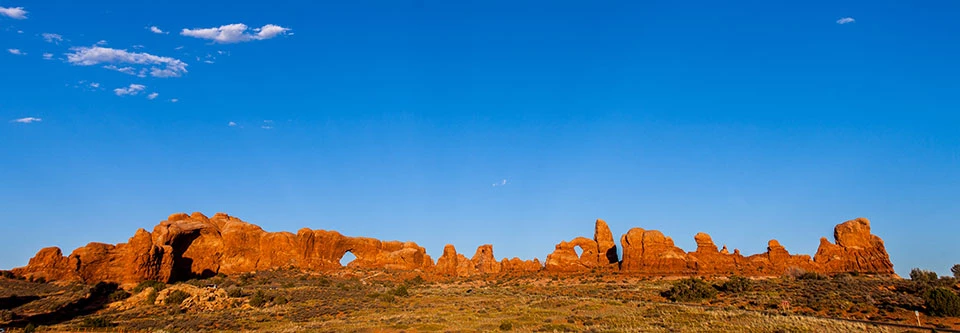 The Windows rock formations from a distance Expansive red rocks in different shapes and sizes, creating a wall. There are multiple arches among the wall and blue sky can be seen through them. The foreground is full of low-lying vegetation and the sky is brilliant blue.
