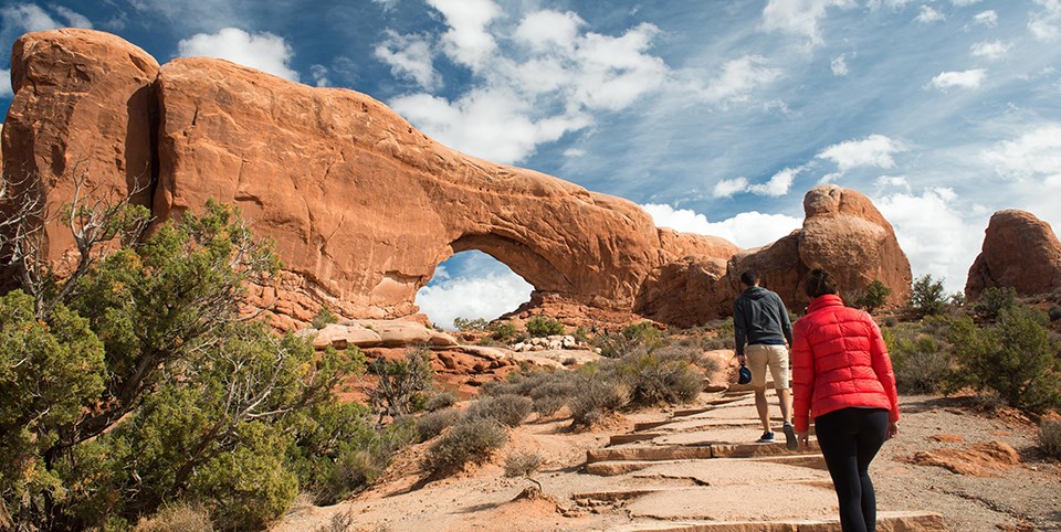 A person in a red jacket hikes behind a person in a gray jacket. The trail they are walking is tan stone with green bushes around them. Up ahead of them is a large red arch with a blue sky with puffy white clouds behind it.