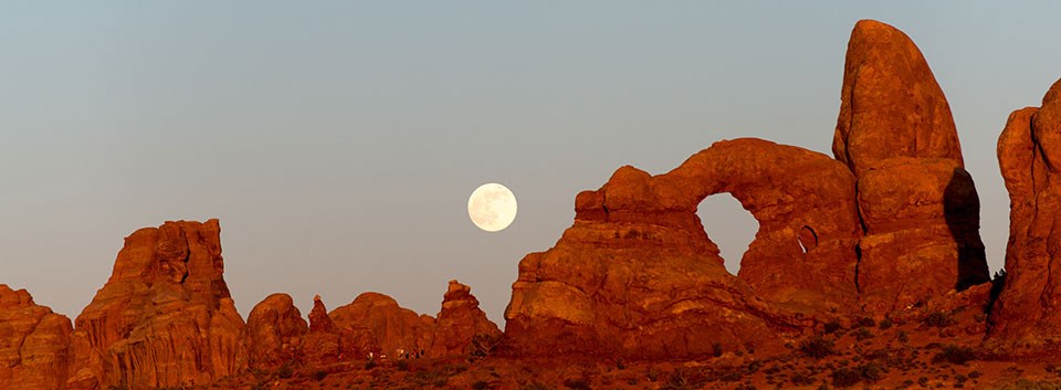 Red rock peaks and spires of varying sizes and shapes with an arch. The full moon rises behind them.