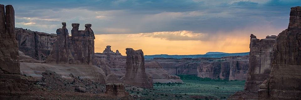 Tall and dramatic red rock pinnacles and towers are backdropped by storm clouds hovering above a golden sky as the sun sets. Between the red rock towers the ground is covered in green vegetation.