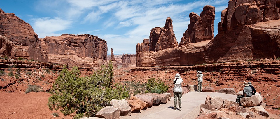 View down a canyon of red rocks with a blue sky with wispy white clouds. In the sandy area below the canyon walls are green shrubs. There is a small paved area where two people stand and two sit together on a large rock.