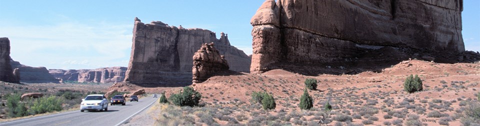 Cars drive on a road that cuts through massive red rock towers.
