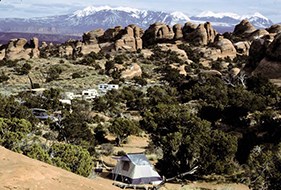 campsites are dotted along an area growing with green trees. In the background are snow capped mountains,