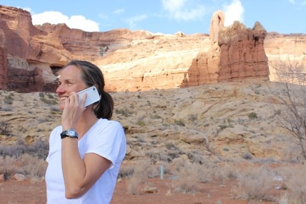 A woman in a white shirt talks on a cell phone in front of a tall red rock walls and towers.
