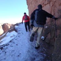 two hikers walk along a snowy and icy trail with a rock wall to one side and a sloping cliff to the other