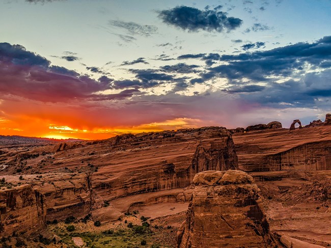 A large wall of orange colored rock with a lone rock arch on top. In the background is a yellow, orange, pink and blue colored sunset with clouds.