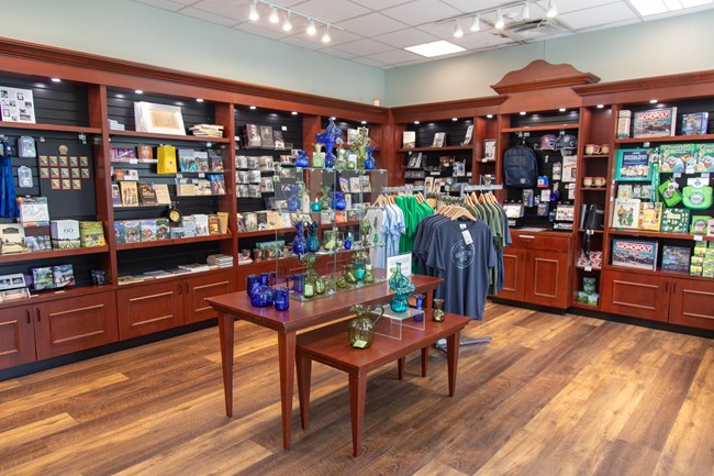 Shelves along a wall with books and toys, and a display in the middle of a room with colored glass vases and t-shirts.