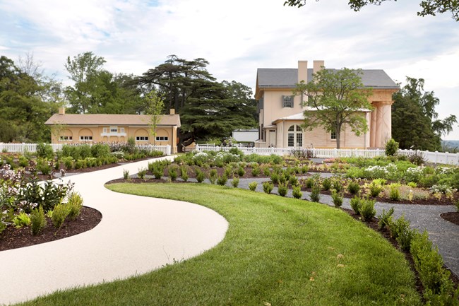 A sidewalk winds through a garden towards two buildings, one three-stories tall with large columns.