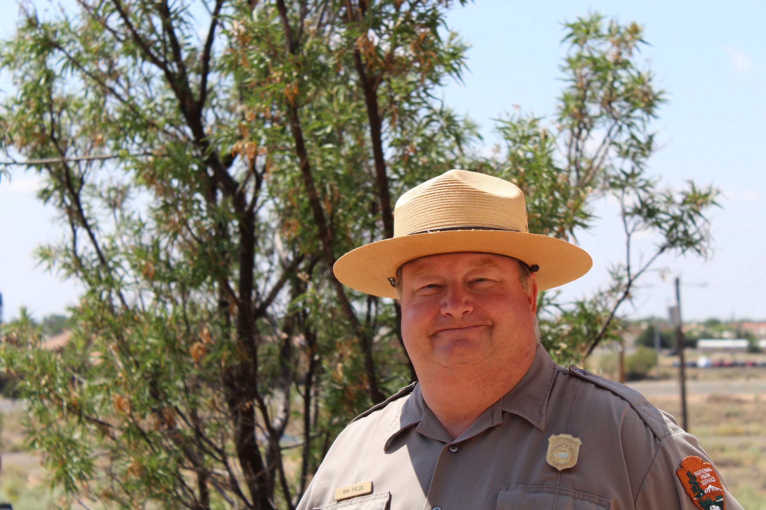 Man in uniform and a hat stand standing in front of a tree.