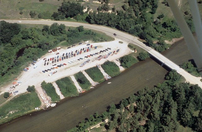 Bird's eye view of the Fort Niobrara Launch Area, six white trails connect a large cleared area to river. Vehicles are parked in the cleared area and to the far right of the photo a bridge crosses the river.