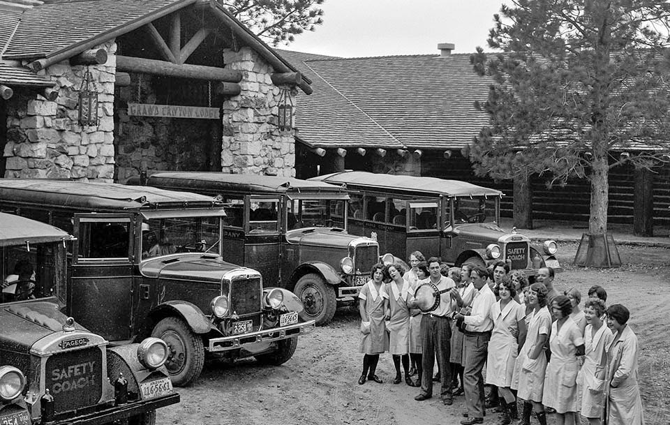 around 20 young lodge employees in uniform are singing to guests sitting in 1930's tour buses that are parked in front of a rustic stone lodge.