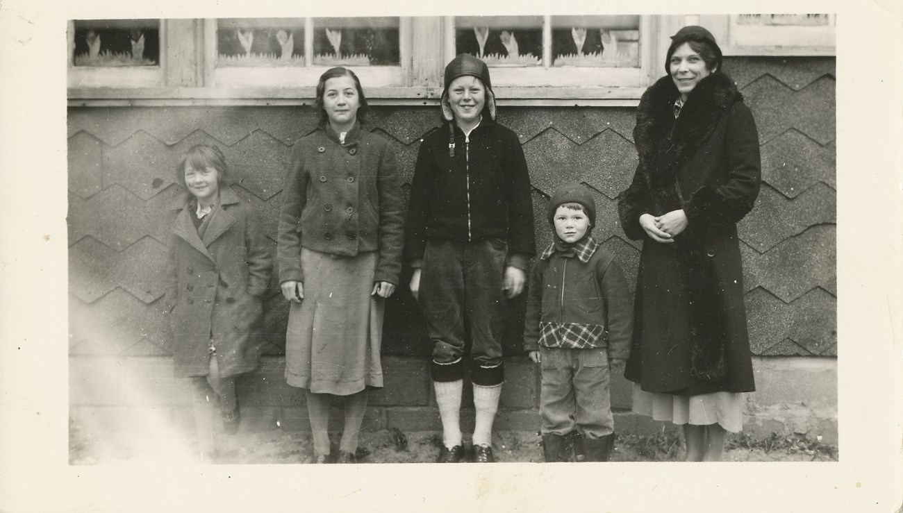 Ronald Rosie standing outside his school with his younger brother, two classmates, and teacher. Four children and an adult standing against a building with zig-zag siding