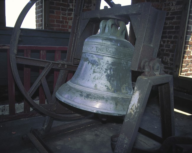 Small light greenish church bell, suspended from metal bar, with circular metal wheel visible
