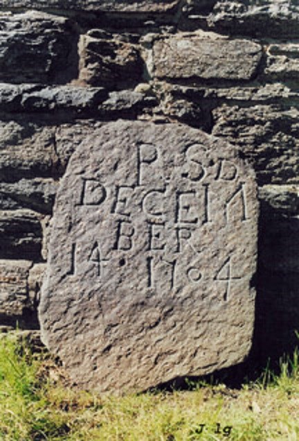 Light grey gravestone, with lettered inscription, leaning against a stone building