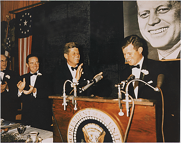 Ted Kennedy stands behind a podium on the right.  He is smiling and standing next to him, President Kennedy claps his hands.
